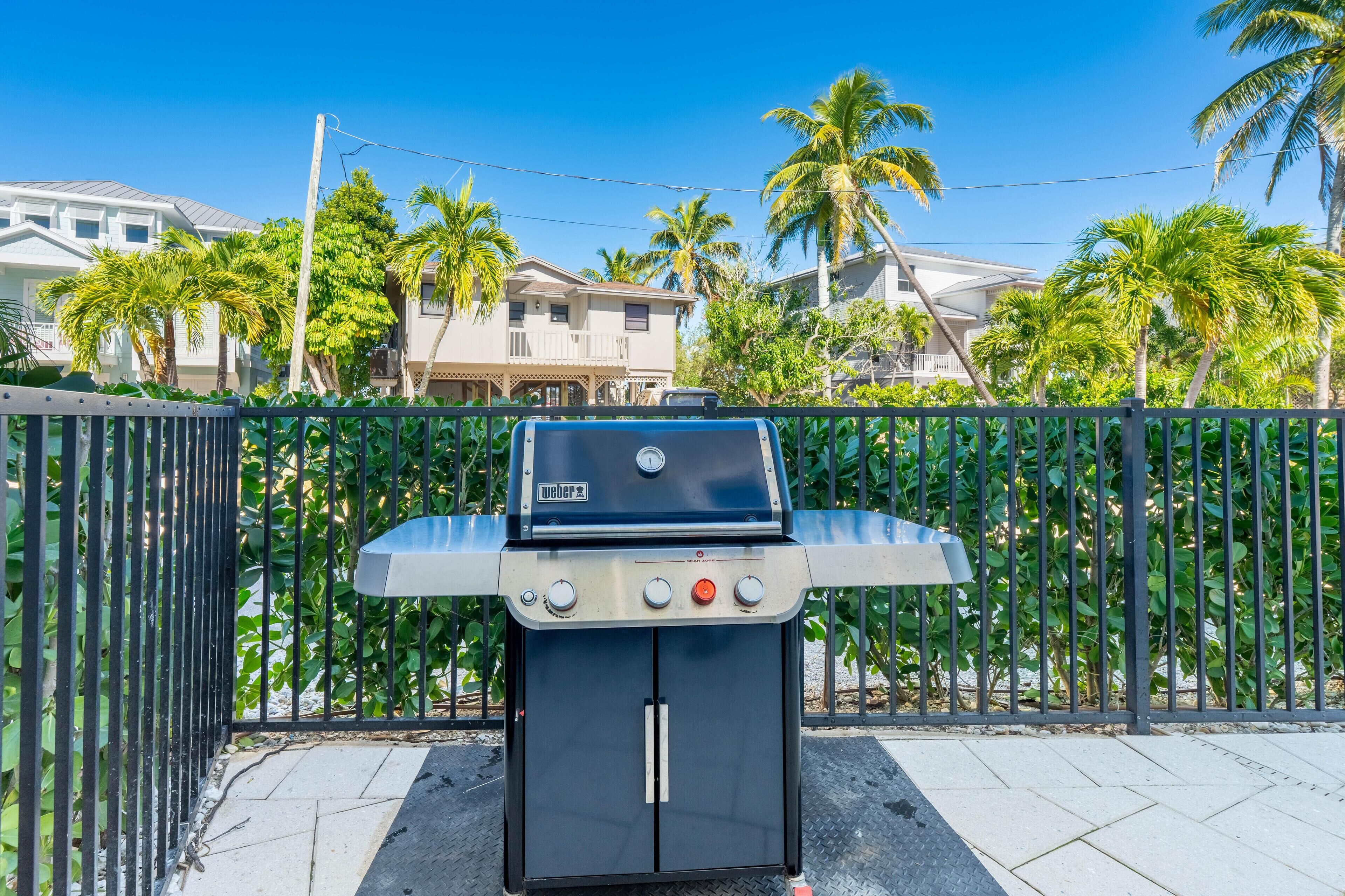 A barbecue grill with a digital display, surrounded by a black metal fence, palm trees, and houses in the background.