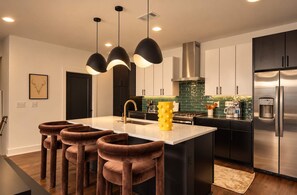 A modern kitchen with a central island, bar stools, stainless steel appliances, and pendant lighting. - Stylish 3BR Nash Escape | Hot Tub + Skyline Views! (Nashville)