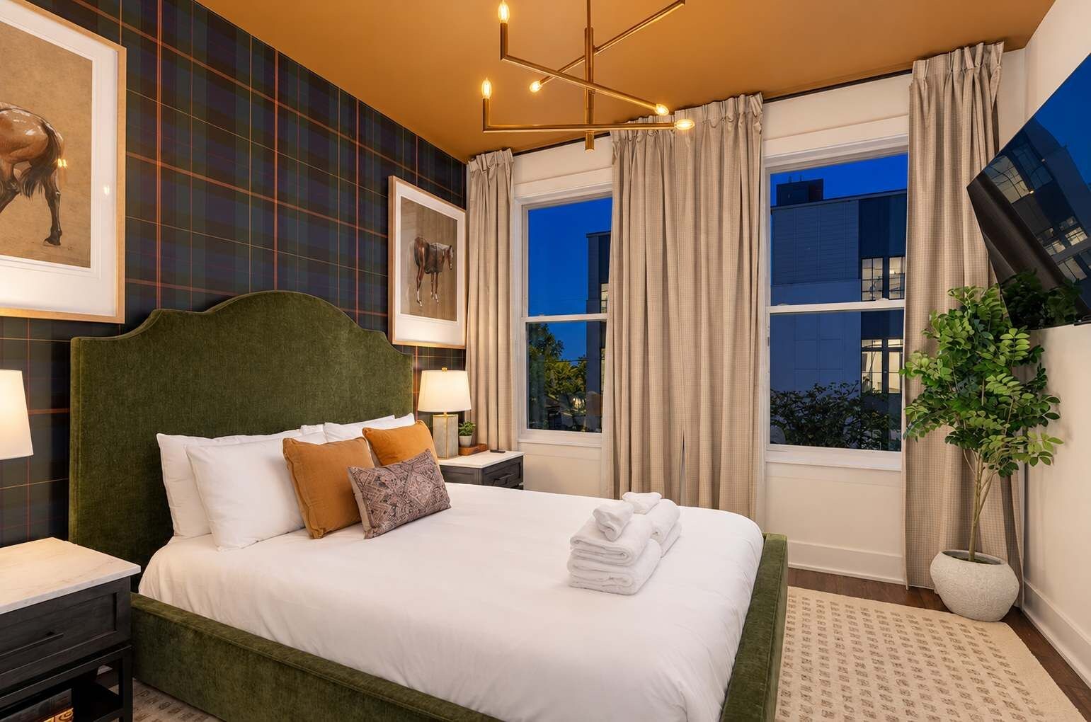 A hotel room with a large bed, a headboard, a nightstand, a lamp, a window with curtains, and a plant.