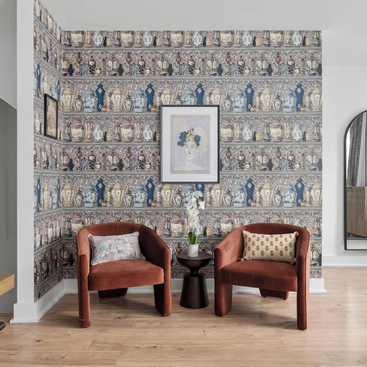 A room with a patterned wallpaper featuring vases and a framed artwork above two brown armchairs with a small table between them.