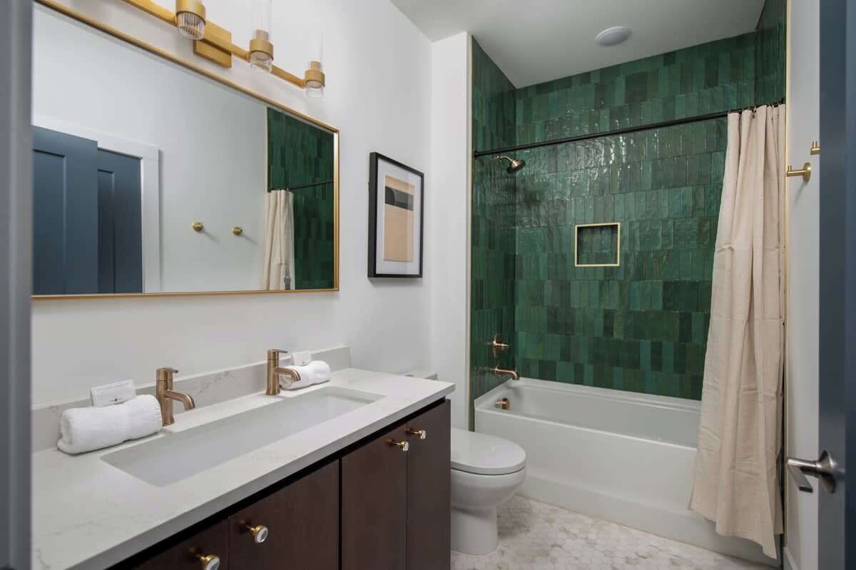 A modern bathroom with a large mirror, two sinks, a toilet, and a shower with a green mosaic wall.