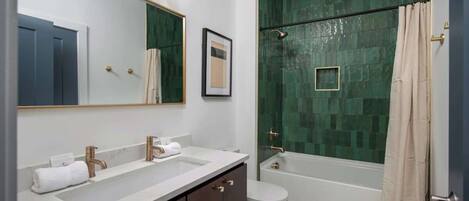 A modern bathroom with a large mirror, two sinks, a toilet, and a shower with a green mosaic wall.