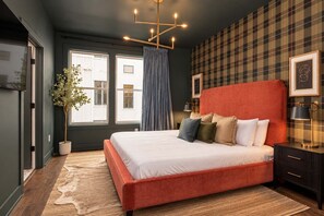 Ein Schlafzimmer mit einem großen Bett, einer Topfpflanze, einem fensterförmigen Öffnung mit Vorhängen, einer karierten Wand und einer modernen Leuchte.