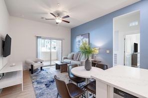 A modern living room with a ceiling fan, a flat-screen TV, a dining area with a white table and chairs, and a large window with a view of buildings. - Landing | Stunning 3BD, Pool (Casas Adobes)