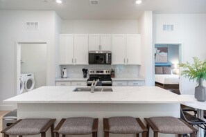 A modern kitchen with white cabinets, a central island with stools, a built-in microwave, and a dishwasher. - Landing | Stunning 3BD, Pool (Casas Adobes)