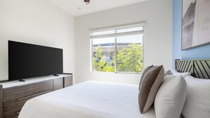 Un dormitorio con una cama, un televisor sobre un mueble, una ventana con persianas y un cuadro en la pared.
