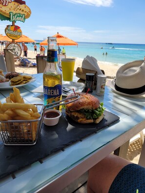 Ein Tisch am Strand mit einem Burger, Pommes, einem Getränk und einem Strandsonnenschirm.