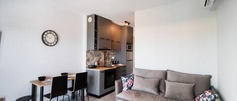 A modern living room with a sofa, a coffee table, and a dining area with chairs and a table. There is a kitchen visible in the background.