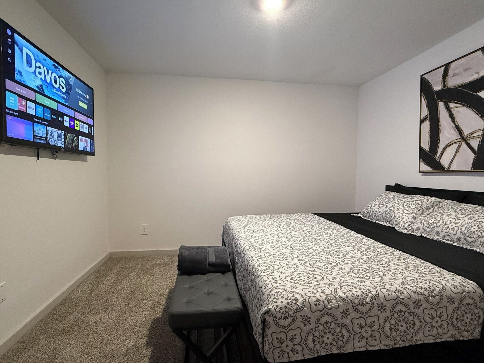 A bedroom with a bed, a bench, a TV mounted on the wall displaying channels, and a large abstract painting on the wall.