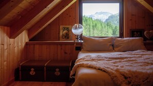 Ein gemütliches Schlafzimmer in einer Holzhütte mit großem Fenster und Blick auf die Berge, einem Bett mit weißer Bettwäsche und einer kleinen Kommode.