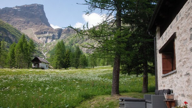 Ein Berggeländehaus mit Steinfassade, einer Terrasse mit Gartenmöbeln und Blick auf ein Tal und Berge im Hintergrund.