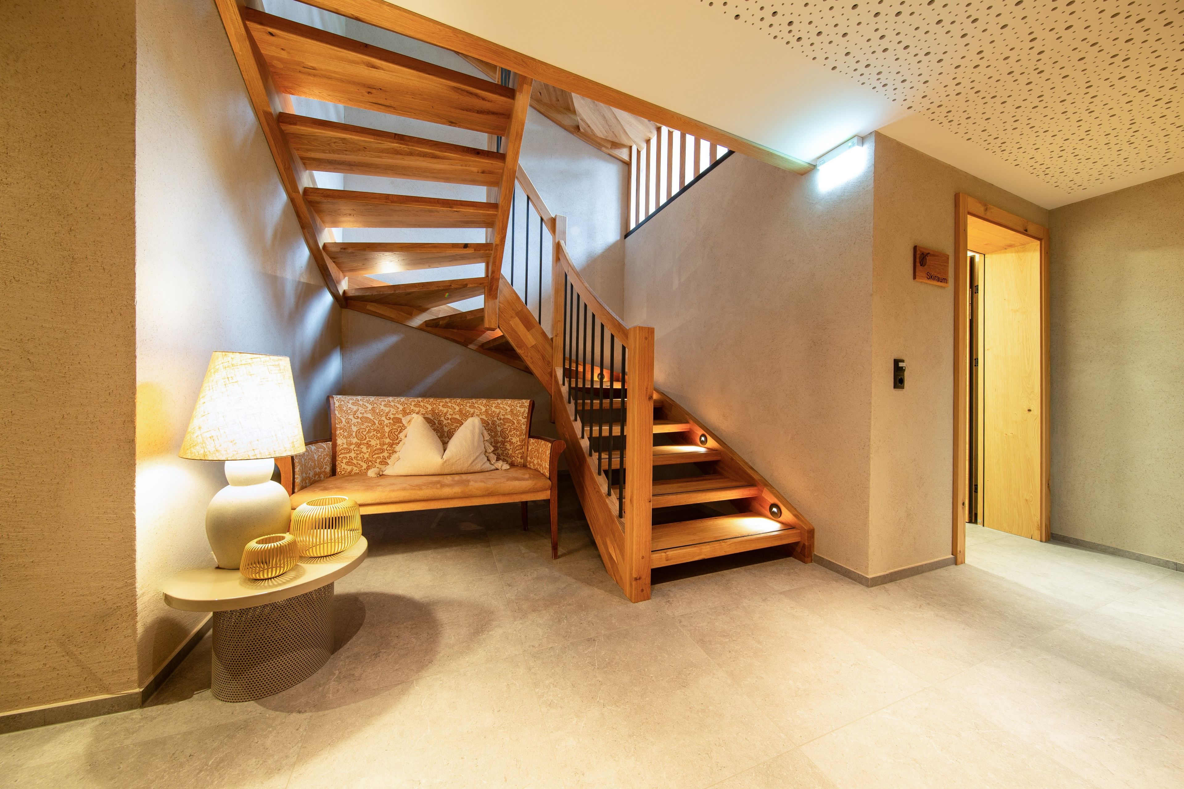 A staircase with wooden railings leading to an upper floor, a sofa with patterned cushions, a small table with a lamp, and a wall-mounted light switch.