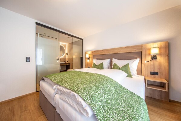 A modern hotel room with a large bed, a wooden headboard, bedside lamps, and a glass shower enclosure.