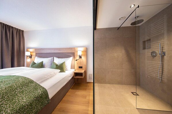 A modern hotel room with a large bed, a bedside table, a nightstand, a glass-enclosed shower, and a wooden headboard.