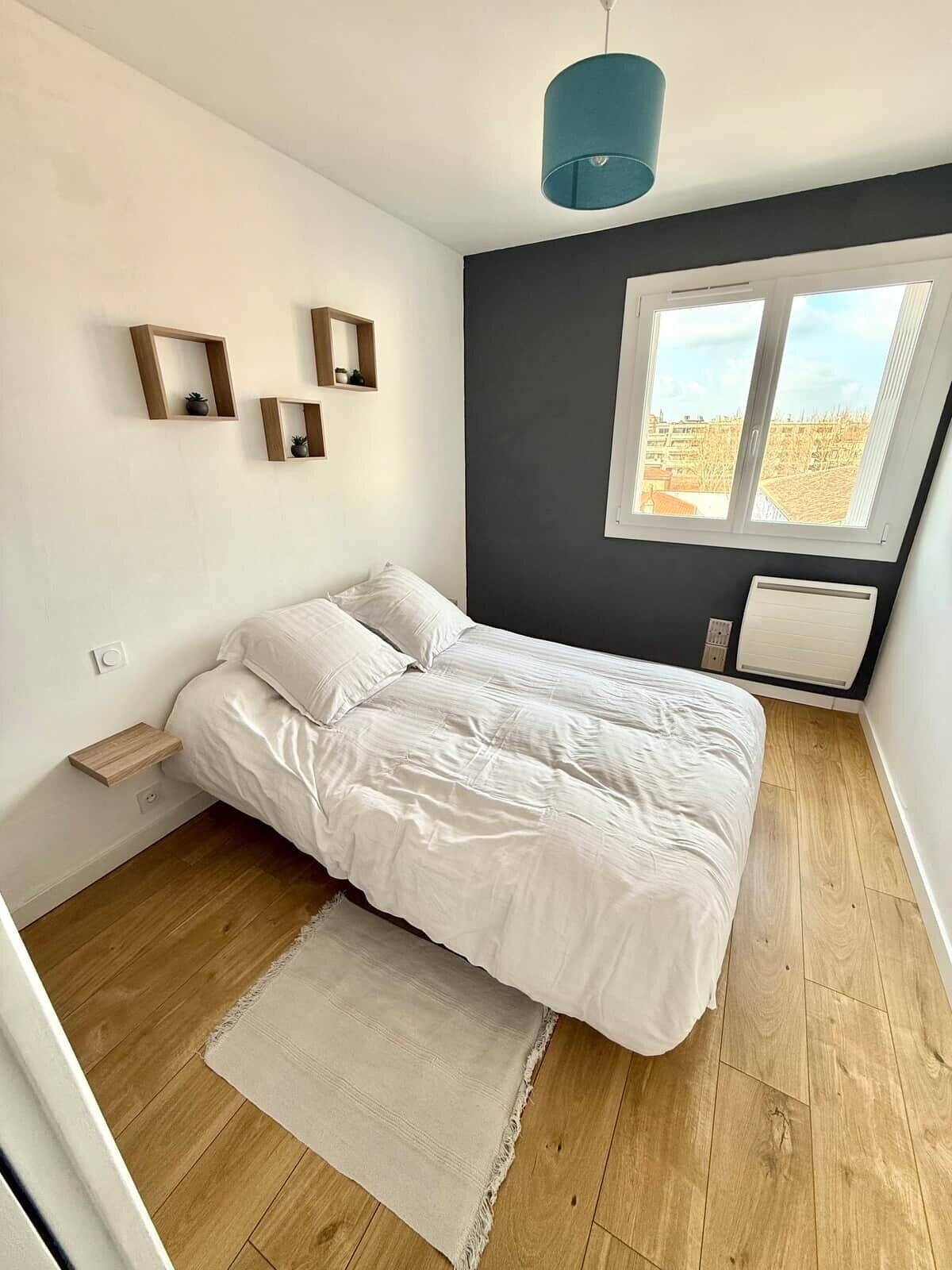A bedroom with a bed, a window, a wall-mounted shelf, and a hanging light fixture.