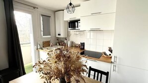 A modern kitchen with white cabinets, a dining table with a vase of dried flowers, and a view of the outdoors. - L'Ecrin St Aubin (Toulouse)