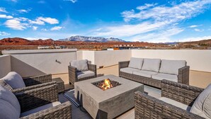 A rooftop patio with wicker furniture and a fire pit, overlooking a mountainous landscape.