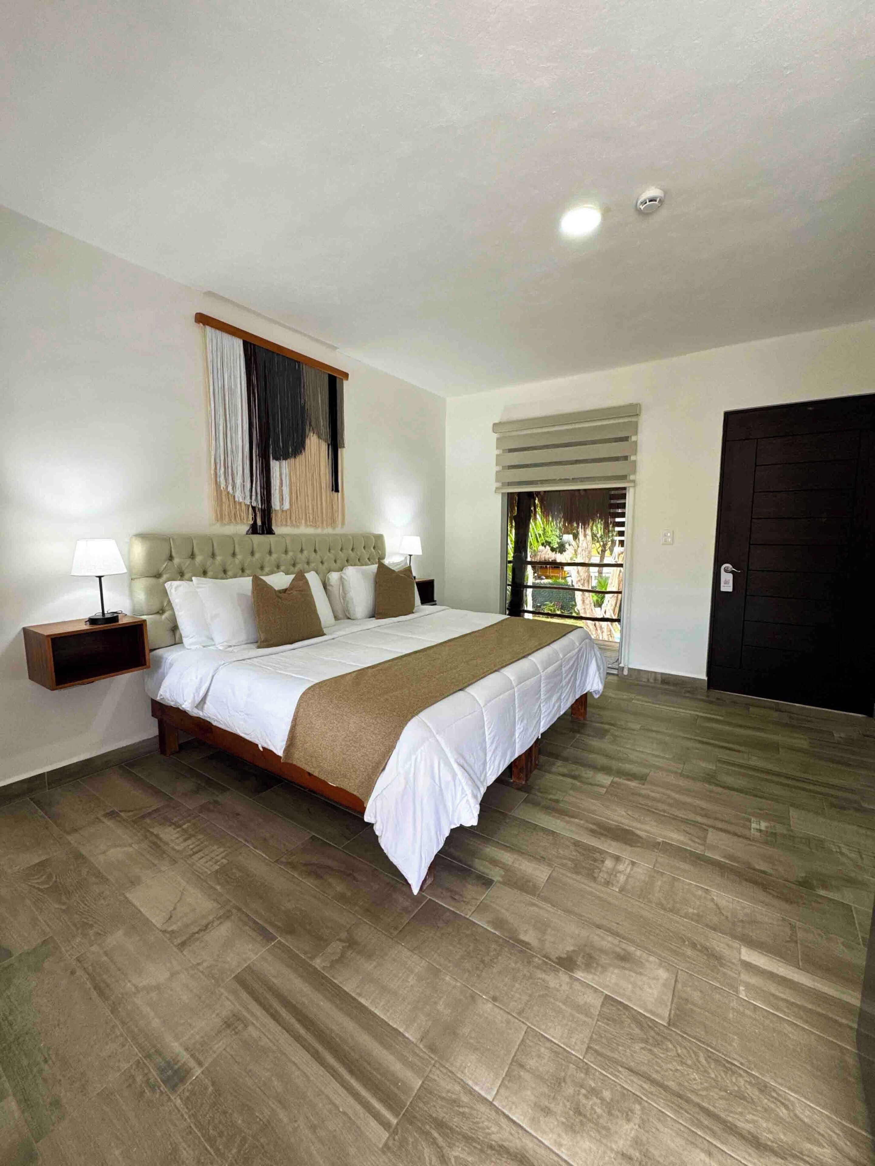 A bedroom with a large bed, two bedside tables, a window with blinds, a door, and a ceiling light.