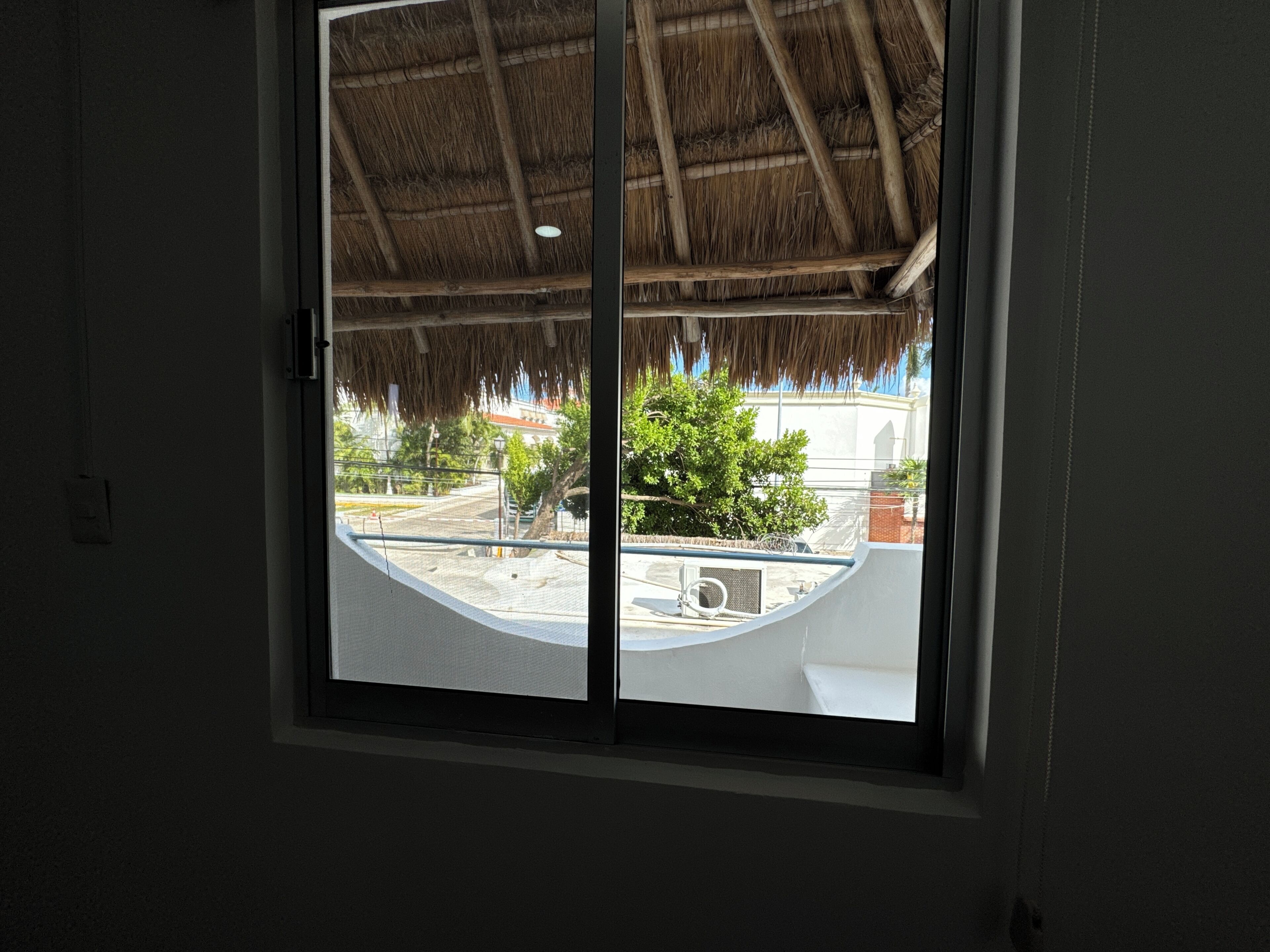A window with a view of a building with a thatched roof, a balcony, and a tree.