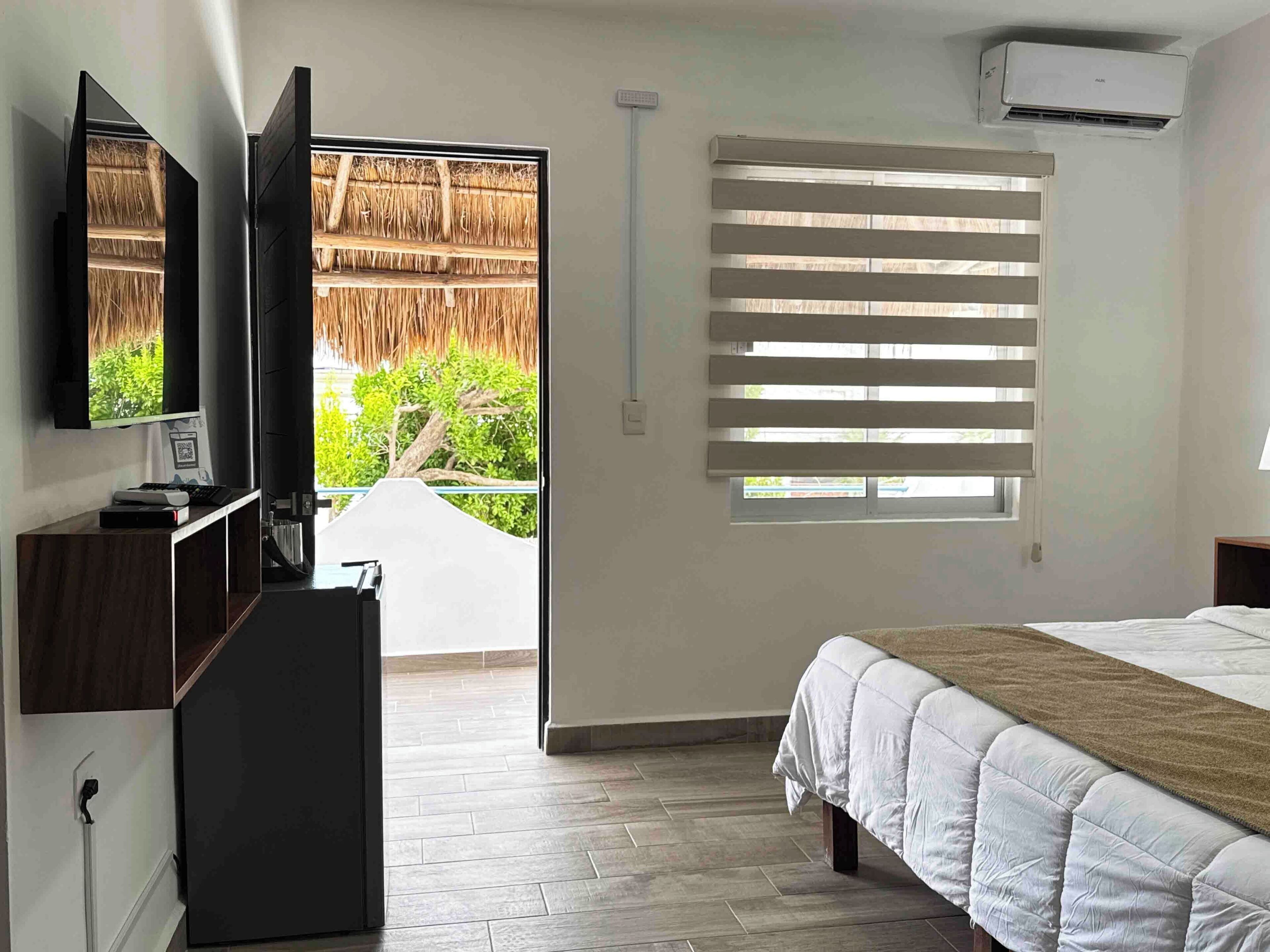 A bedroom with a bed, a TV, a desk, a window with blinds, and an air conditioning unit.