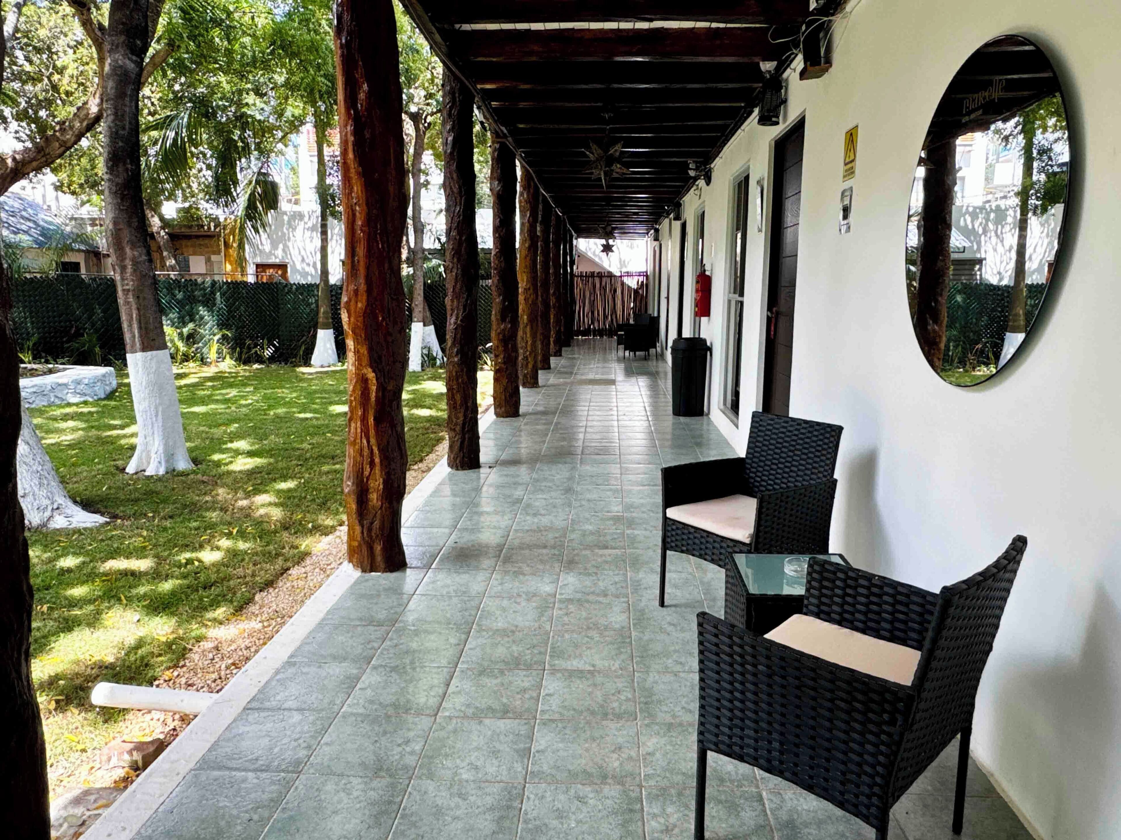 Un espacio al aire libre cubierto con piso de baldosas, mobiliario de mimbre negro y una pared blanca con un espejo circular.