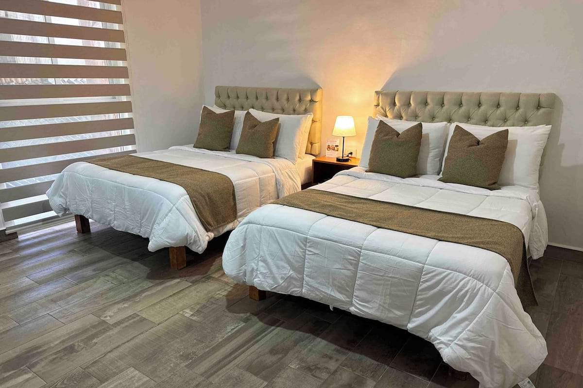 Two twin beds with headboards, a bedside table with a lamp, and a window with blinds.