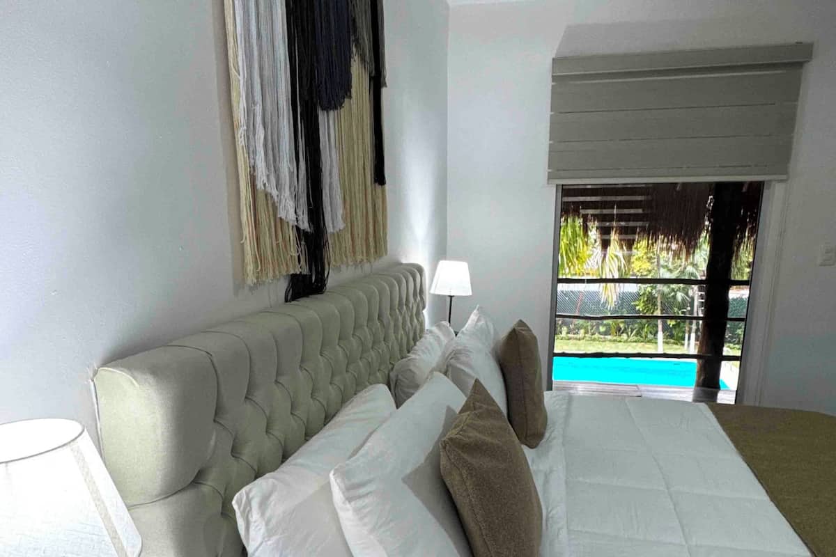 A bedroom with a bed, bedside lamps, a wooden nightstand, and a view of a pool.