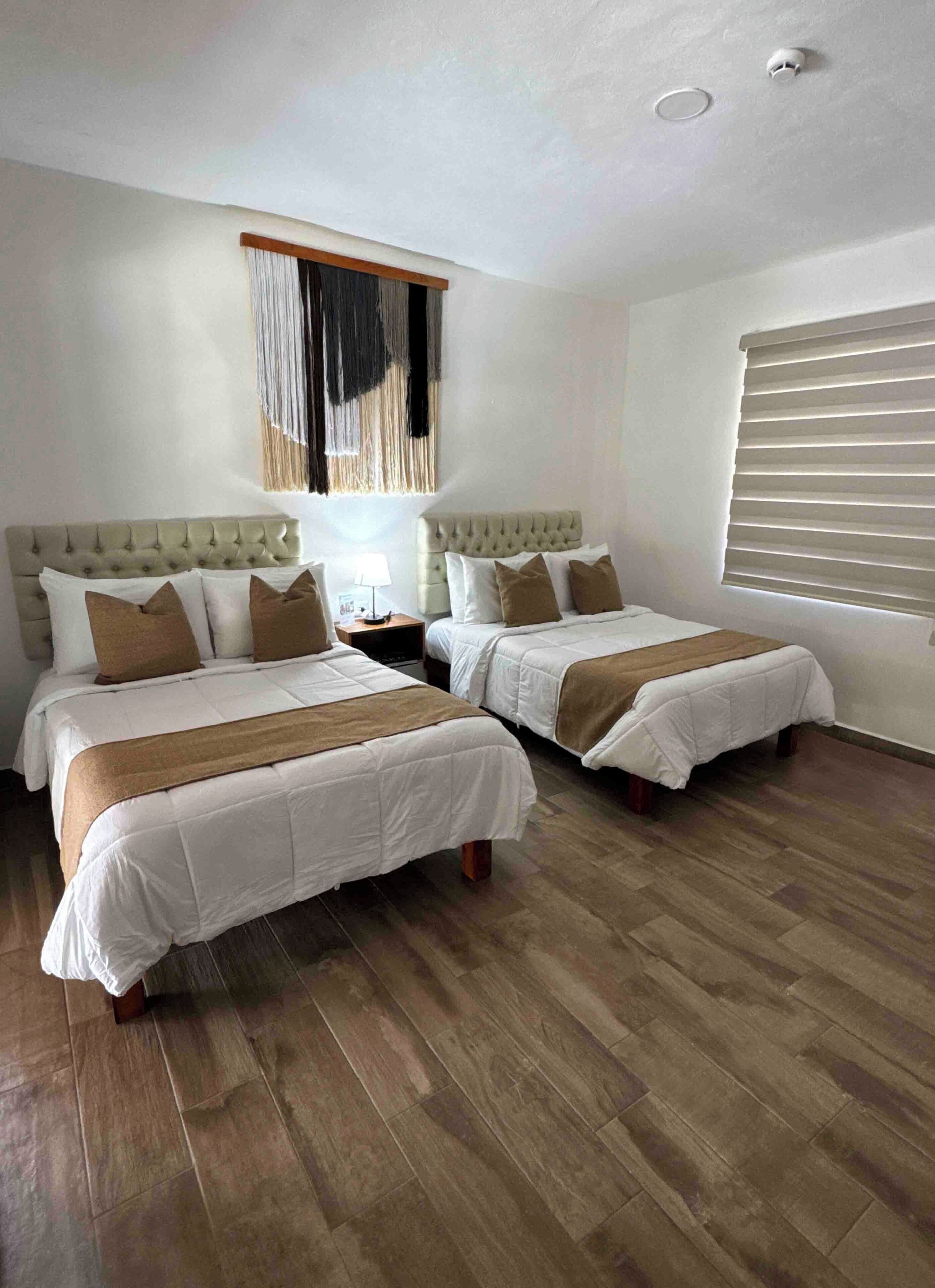 A hotel room with two beds, wooden flooring, a ceiling light, and a window with blinds.