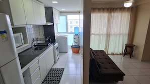 Cocina compacta con gabinetes blancos, un microondas, un fregadero y un refrigerador pequeño. Se aprecia una ventana con cortinas y un ventilador de techo.