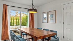 Een eetgedeelte met een houten tafel en blauwe stoelen, een hangende lamp en uitzicht op een tuin met bomen en een gebouw.