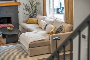 Een gezellige woonkamer met een beige bank, een klein bijzettafeltje, een open haard en een groot raam met gordijnen.