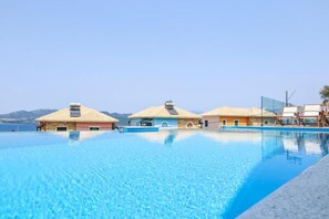 A clear blue infinity pool with three beachside villas, each with a tiled roof and a small balcony. - Red Bee Apartment Sea View (Nikiana)