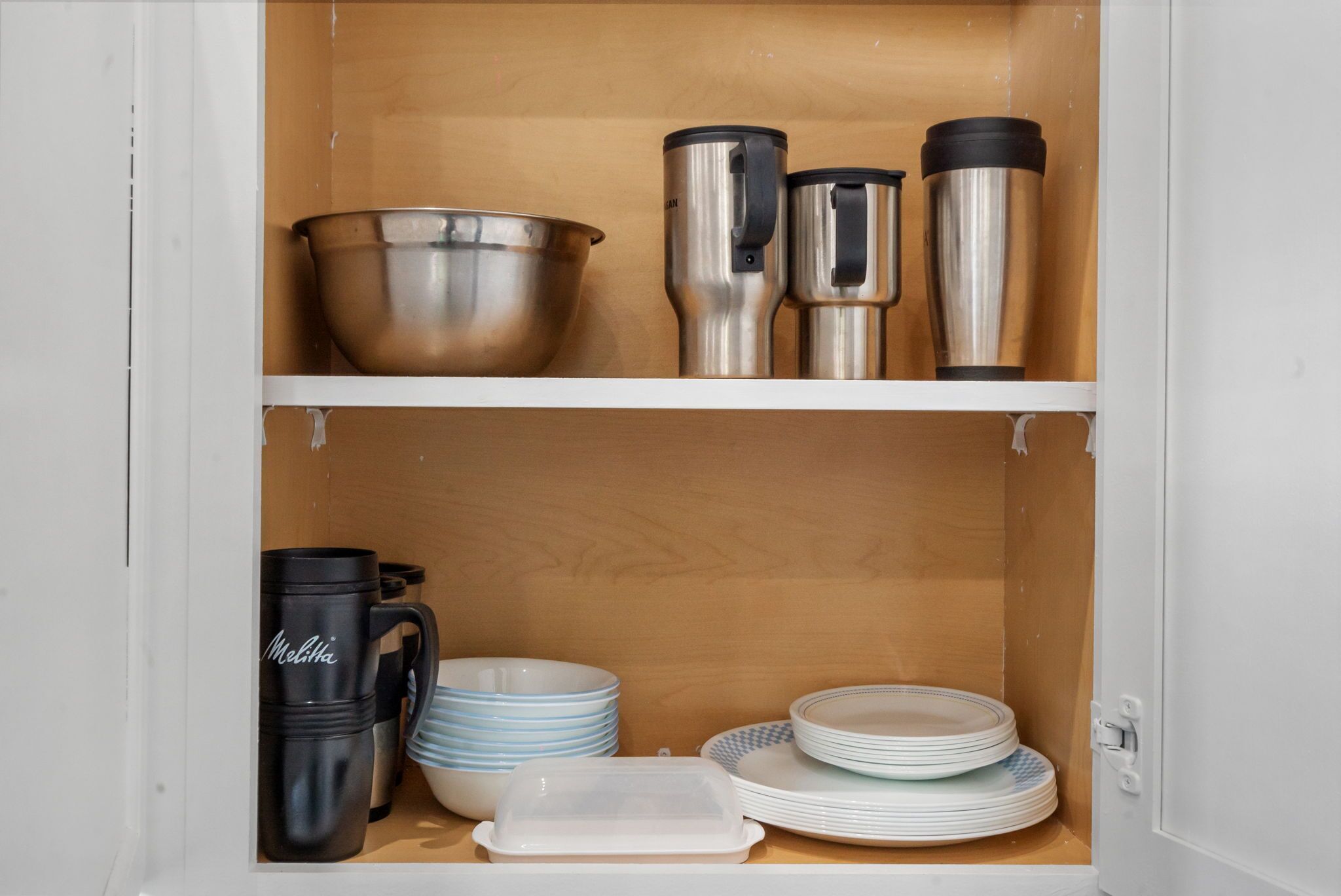 Un meuble de cuisine contenant des ustensiles de cuisine en acier inoxydable, une cafetière noire de la marque Melitta et des assiettes blanches et bleues empilées.