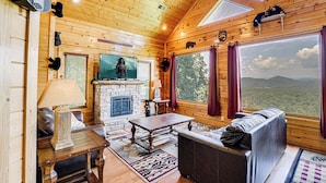 L’intérieur d’un chalet accueillant, avec une cheminée, des murs en bois, un canapé, une table et une grande fenêtre donnant sur une vue imprenable sur les montagnes.