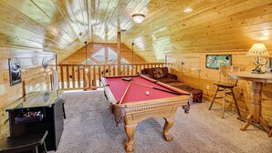Une pièce chaleureuse, avec des murs en bois, comprenant une table de billard, un canapé et une petite table avec une lampe.