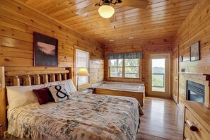 A wooden cabin bedroom with a bed, a nightstand, a window, a bathroom with a bathtub, and a fireplace. - Modern Retreat w/ Stunning Views & Hot Tub (Sevierville)
