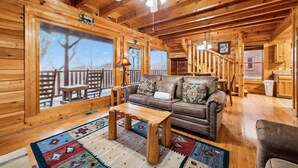 Ein gemütliches Blockhaus-Interieur mit Holzbalkendecke, Deckenventilator, einer Couch mit Kissen, einem Couchtisch und Blick durch große Fenster.