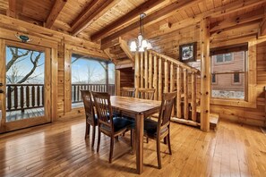 Interior de una cabaña de madera con mesa de comedor, sillas, una escalera y ventanas amplias.
