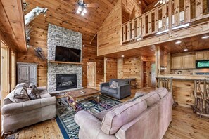 A spacious living area with a stone fireplace, wooden walls, a television mounted above it, and a balcony with wooden railings. - Amazing Smoky Mountain Views w/ Hot Tub (Sevierville)