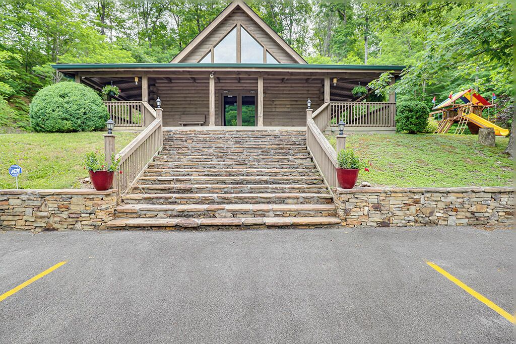 A wooden lodge with a stone staircase leading to a porch, surrounded by greenery and a playground.