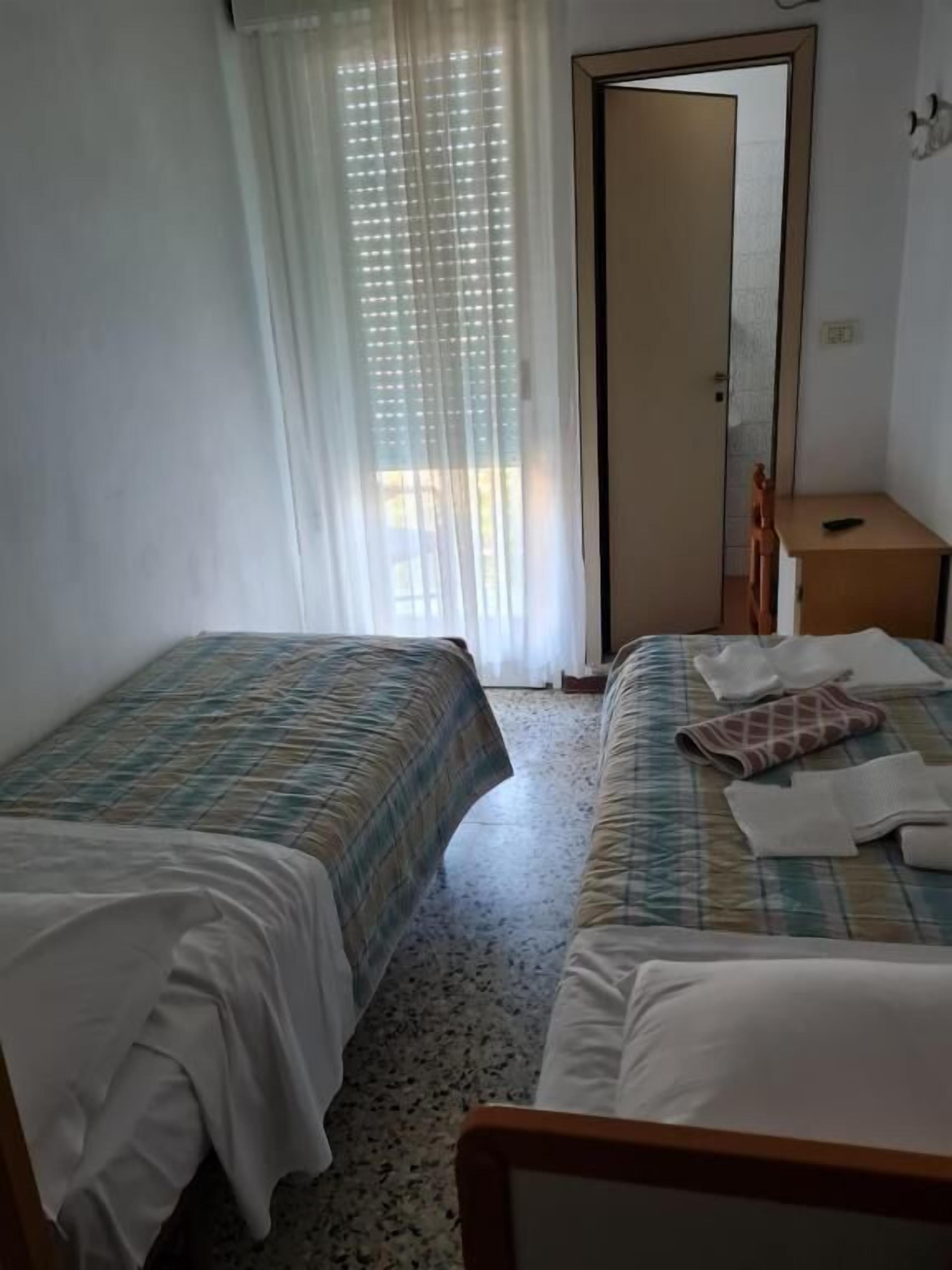 Habitación de hotel con dos camas, una mesita de noche de madera, un espejo y una ventana con cortinas traslúcidas.
