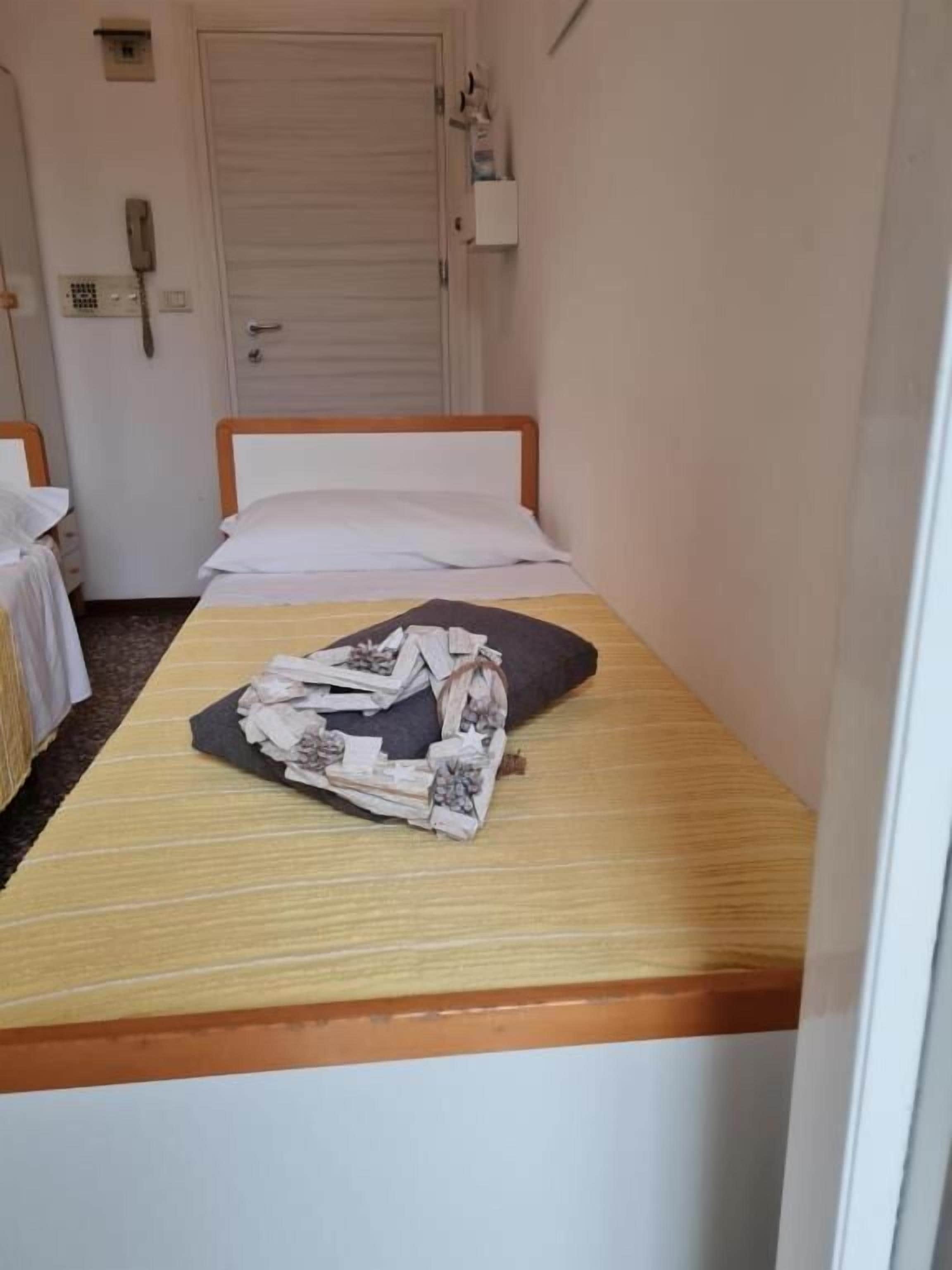 A small, single-bed room with a wooden headboard and a plain white mattress. There is a small bedside table with a lamp and a telephone. The room has a simple, functional design with a door and a small shelf above it.