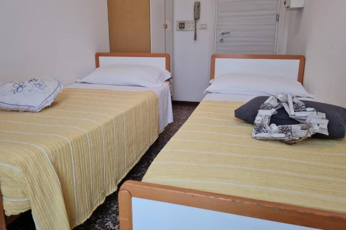 Een kamer met twee eenpersoonsbedden, elk met een houten frame en een gestreept dekbed. Er is een telefoon aan de muur bevestigd en een gesloten deur op de achtergrond.