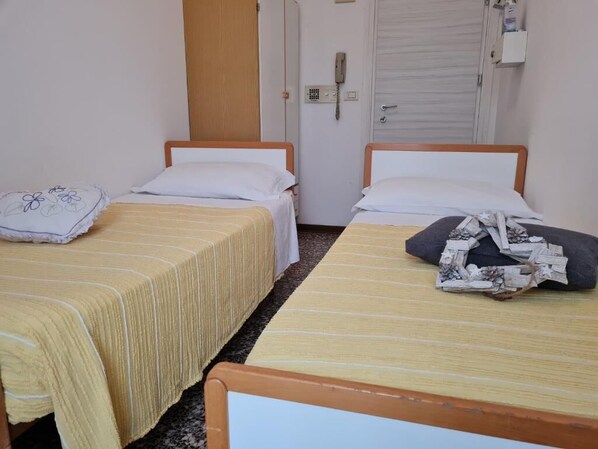 Una habitación con dos camas individuales, cada una con estructura de madera y cubrecama a rayas. Hay un teléfono sujeto a la pared y una puerta cerrada al fondo.