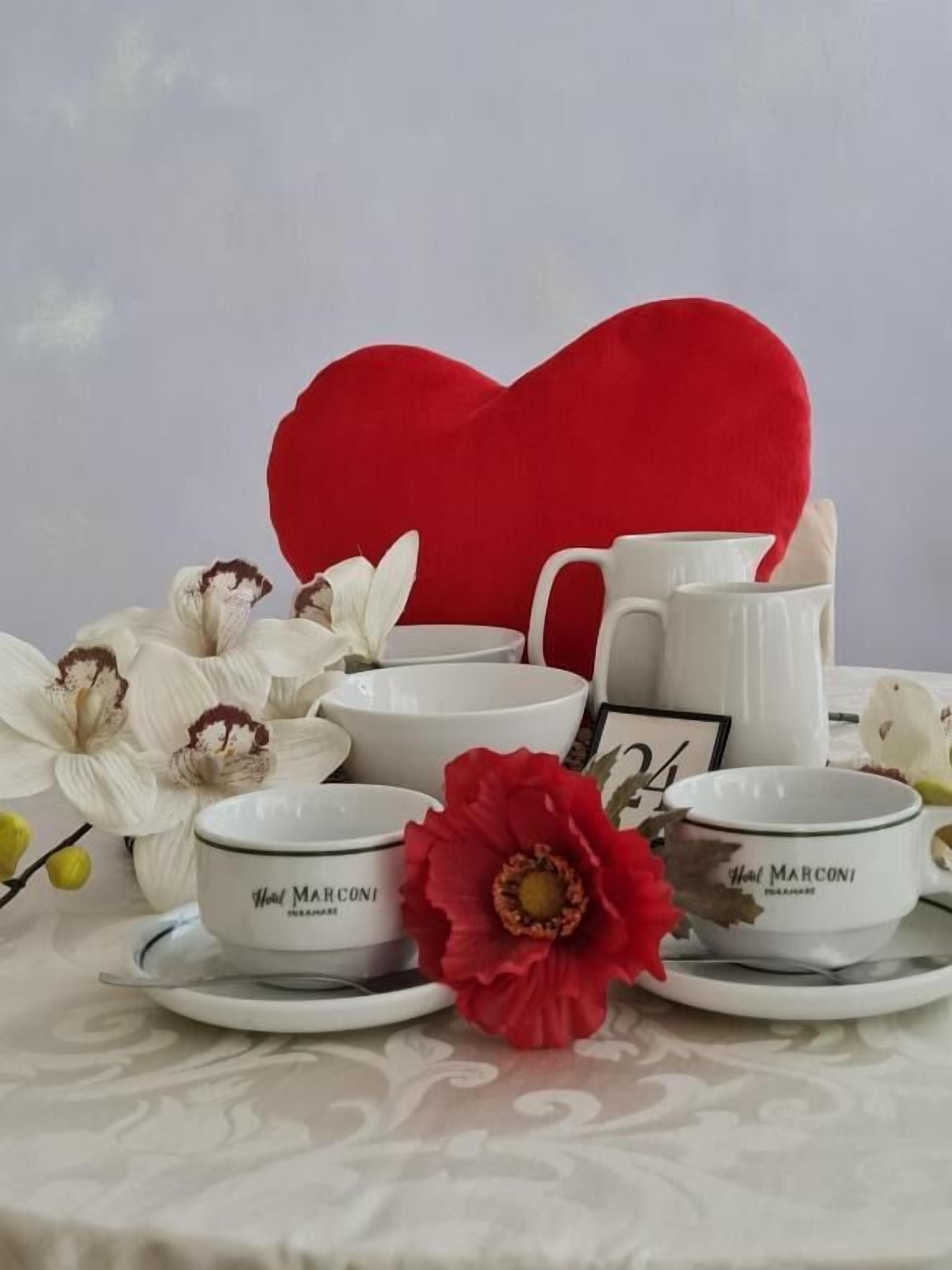 A table setting with white ceramic dishes, a red heart-shaped cushion, and white flowers.