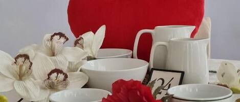 A table setting with white ceramic dishes, a red heart-shaped cushion, and white flowers.