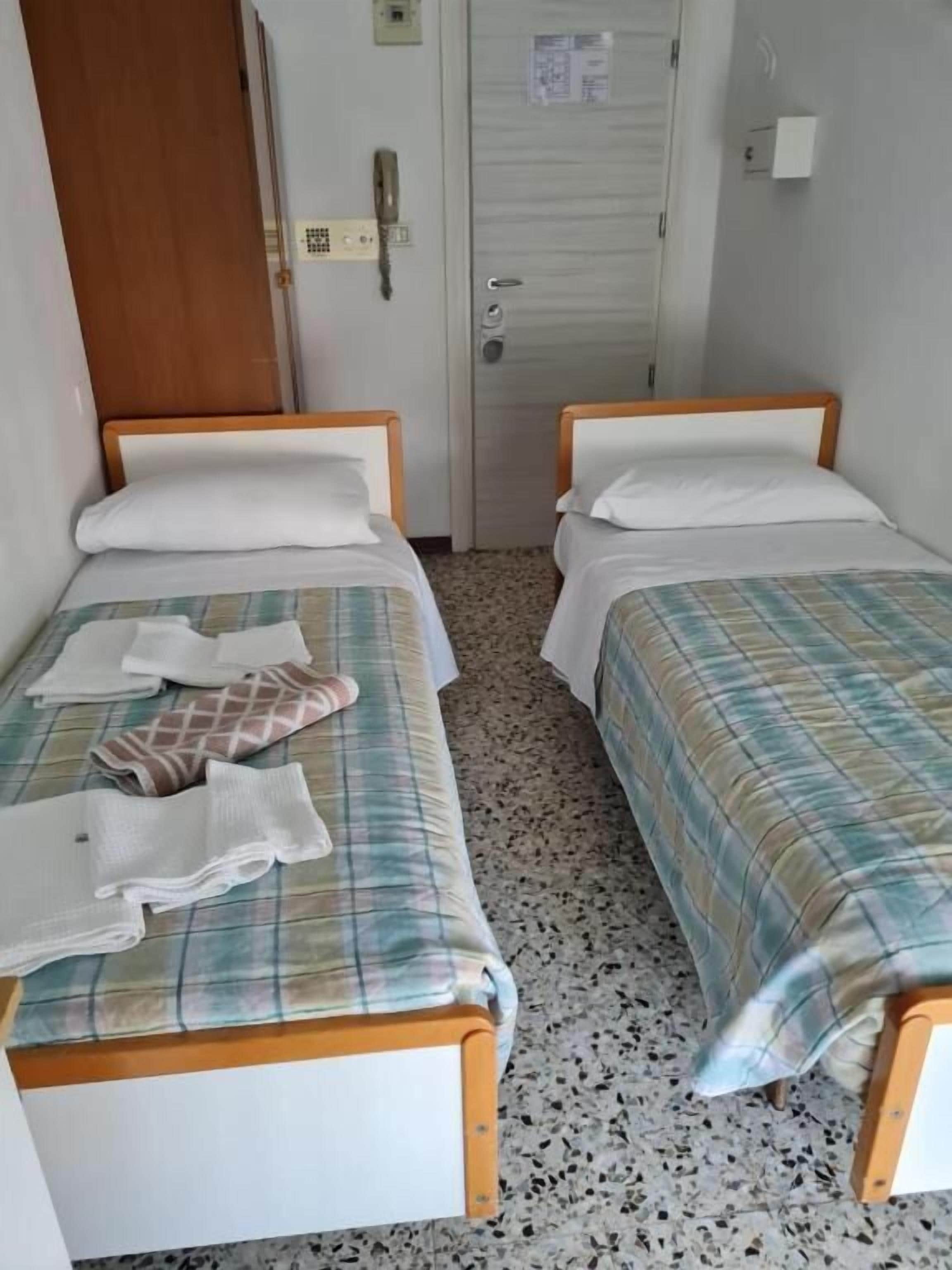 Dos camas individuales con cabeceros de madera y ropa de cama blanca, una mesita con una toalla doblada y una puerta con interruptor de luz y cerradura.