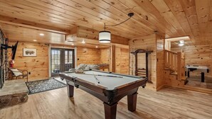 Un intérieur en bois avec une table de billard, une cheminée en pierre et un tableau encadré au mur.