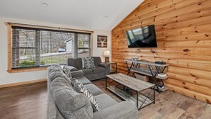 Un salon moderne avec un canapé d’angle, une table basse, un téléviseur fixé à un mur en bois et une fenêtre équipée de stores.