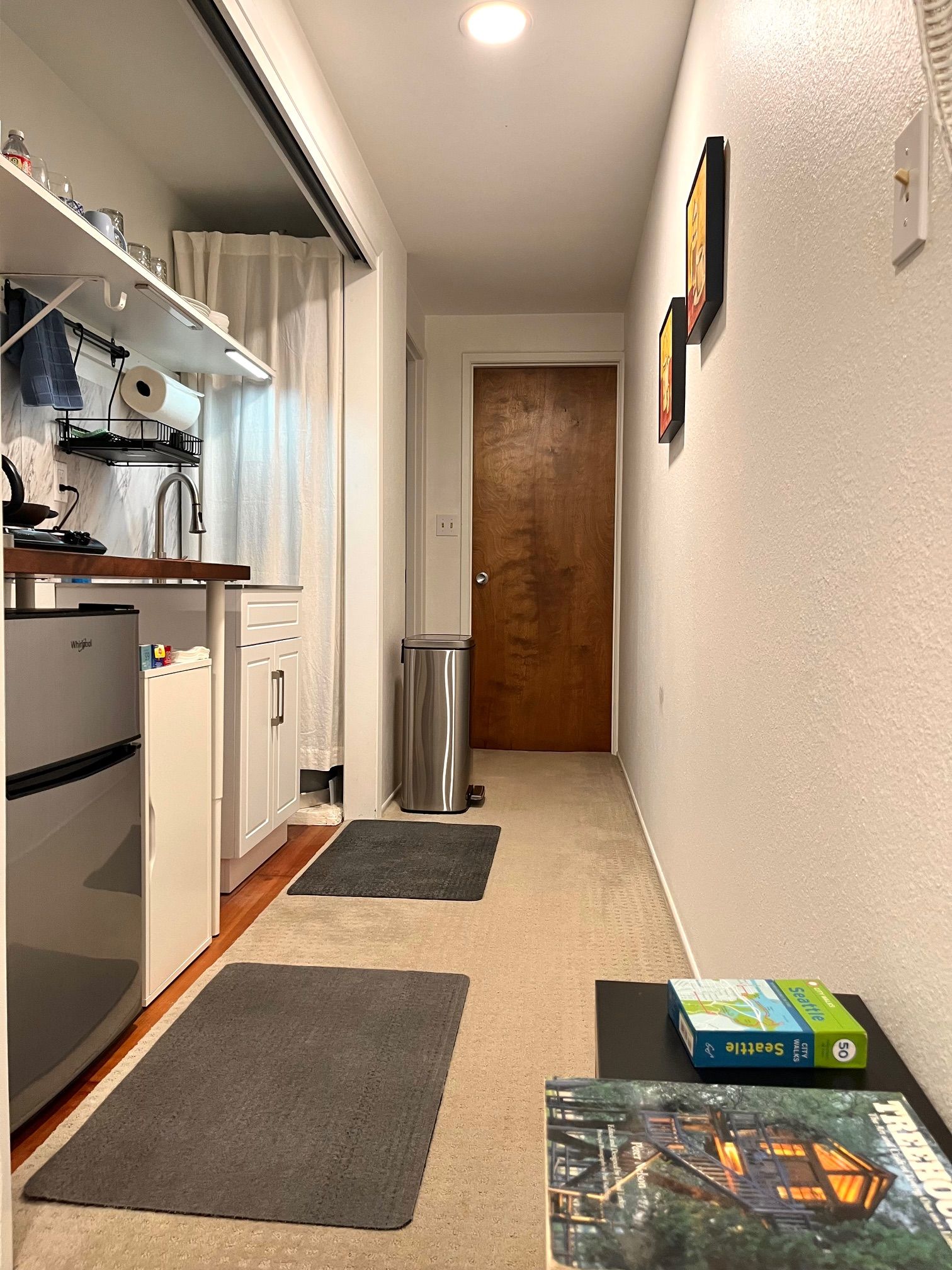 A narrow hallway with a kitchen on one side, a wooden door at the end, and a trash bin in the middle.